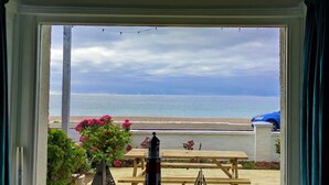 A view from a window overlooking a beach with a wooden table and chairs, a flower pot, and a blue car parked outside. - Absolute Beachfront With Decked Garden Oasis and Views (Bognor Regis)