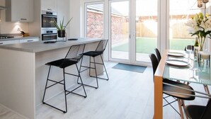 Et moderne køkken med hvide skabe, en central ø med barstole og et spiseområde med et glasbord og stole.
