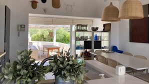 A modern kitchen with white cabinetry, a dining area with wicker chairs, and a view of greenery outside.