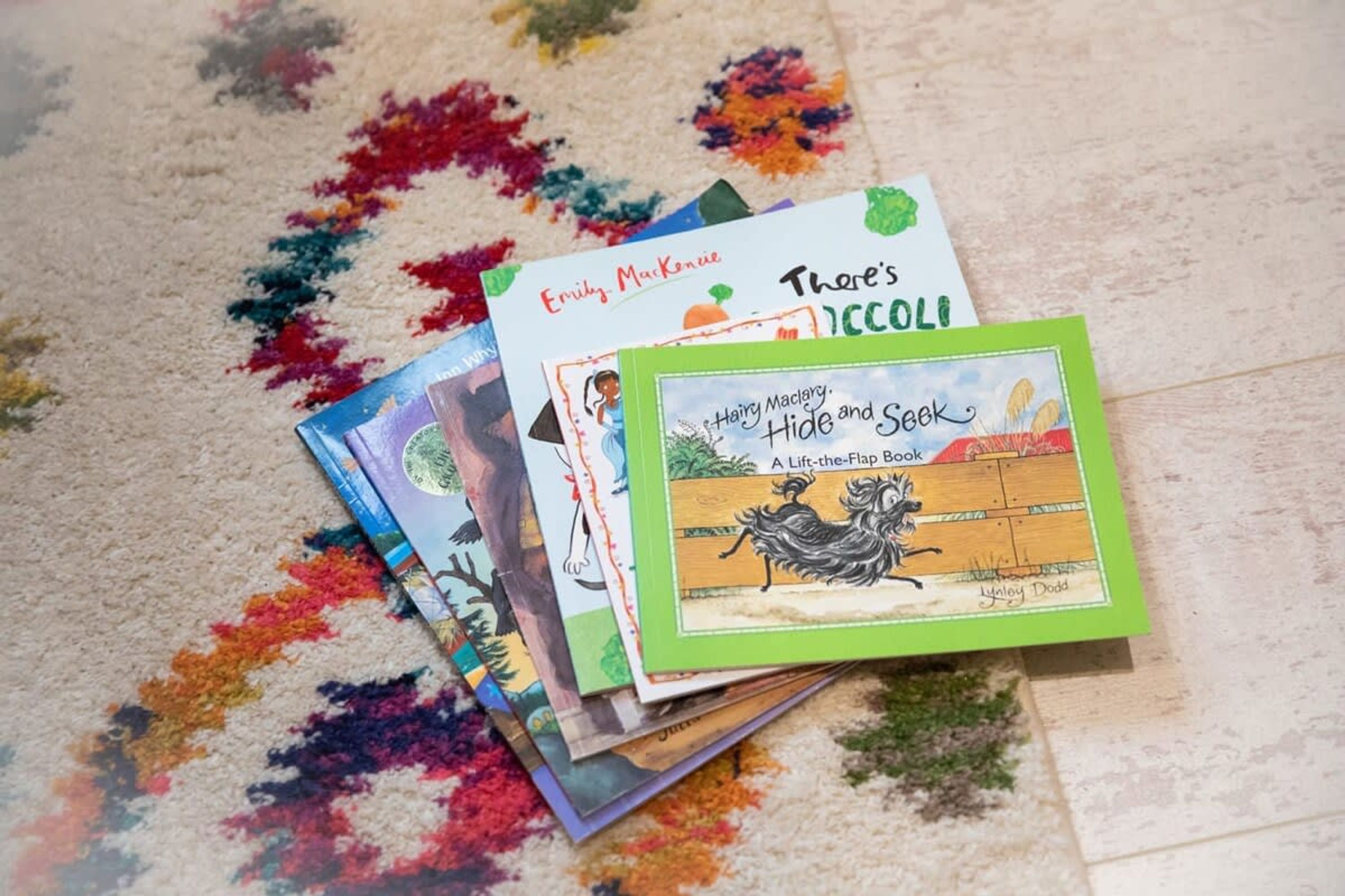 A collection of children's books on a carpet with colorful stains.