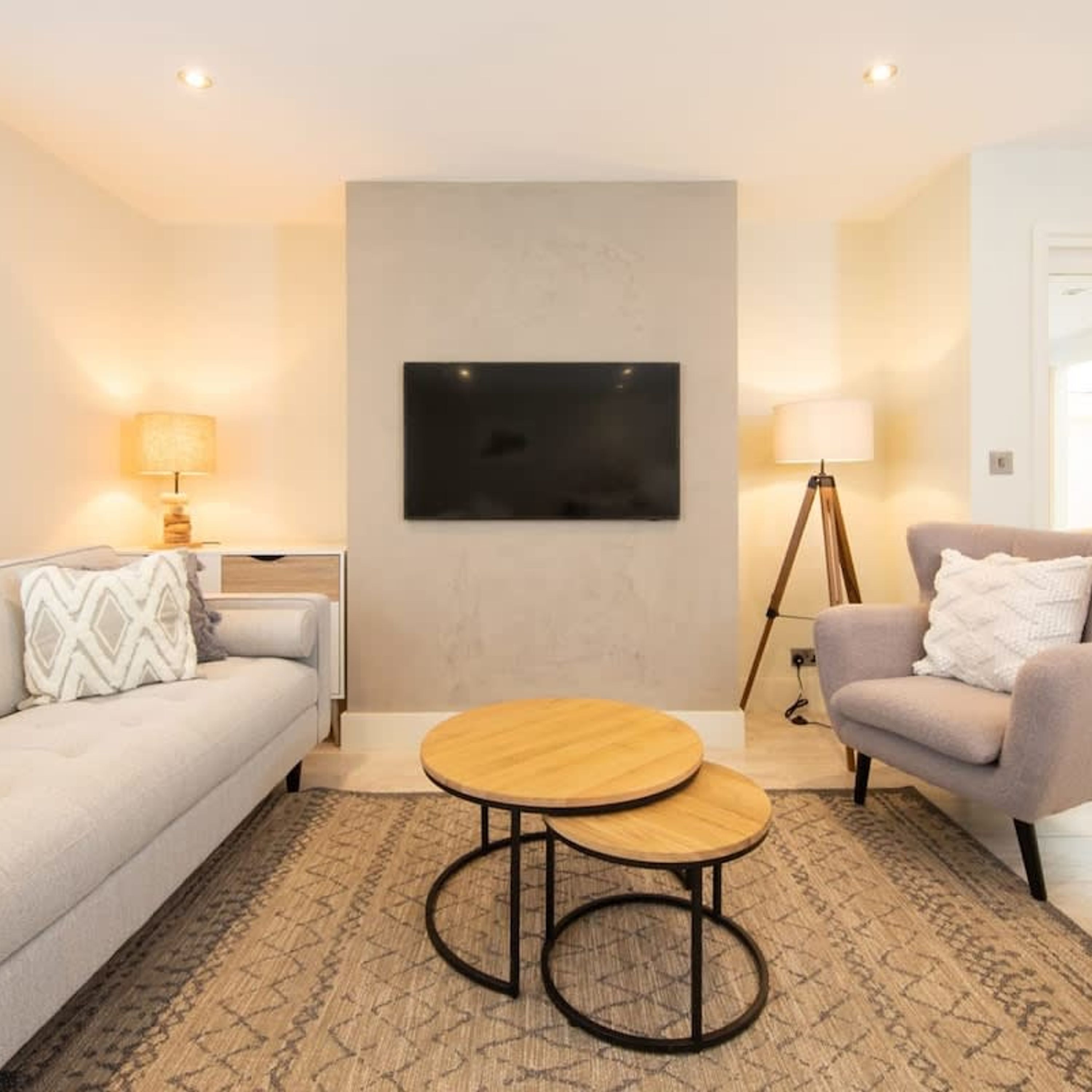 A modern living room with a sofa, armchair, round wooden coffee table, and a flat-screen TV mounted on the wall.