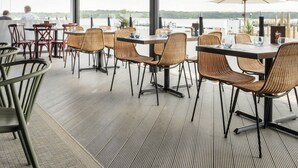 Ein Essbereich im Freien mit Korbstühlen und Tischen, großen Fenstern mit Blick aufs Wasser und einer überdachten Terrasse.