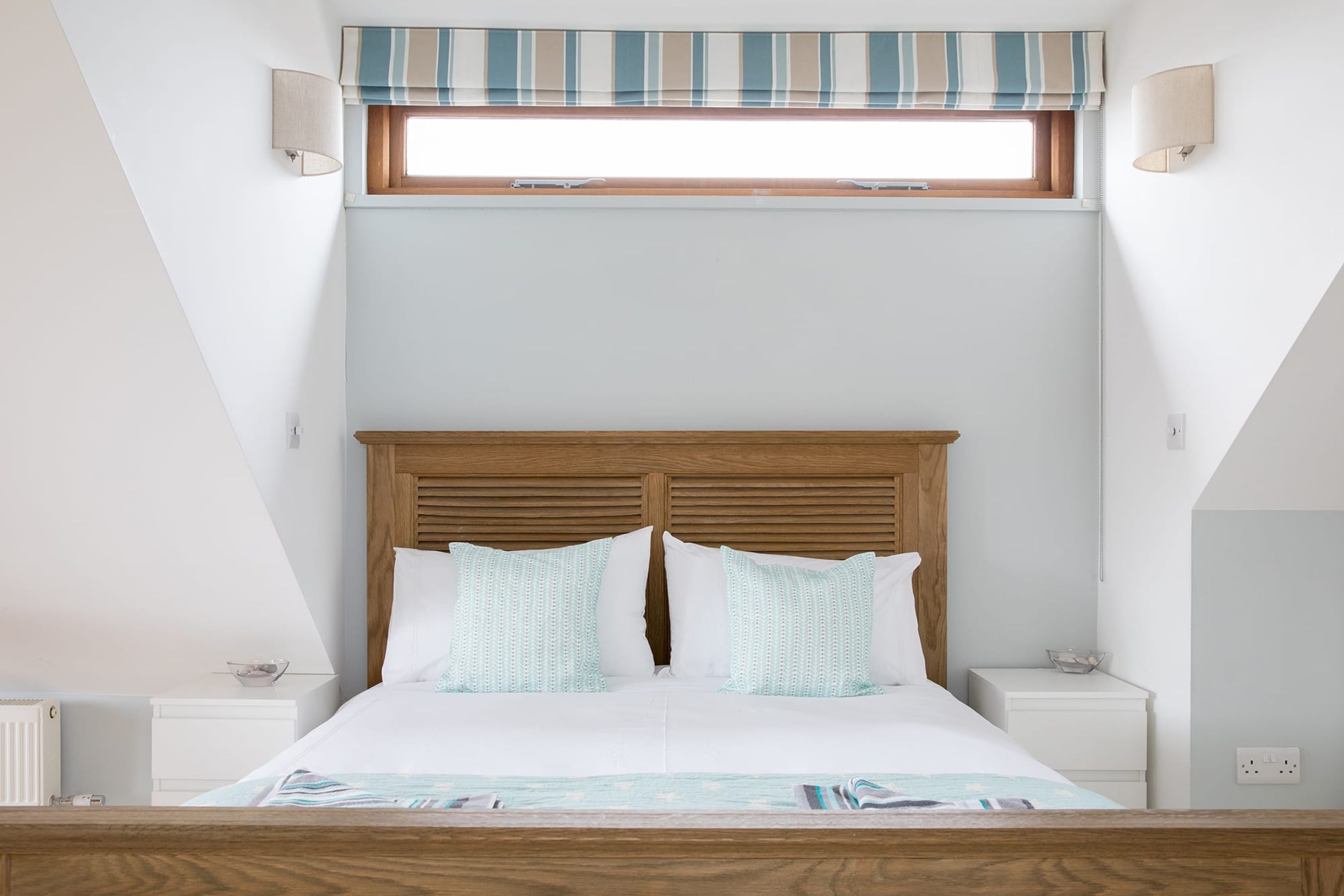 Quarto com cabeceira de madeira, duas mesas de cabeceira, janela com cortina listrada e cama com roupa de cama branca.