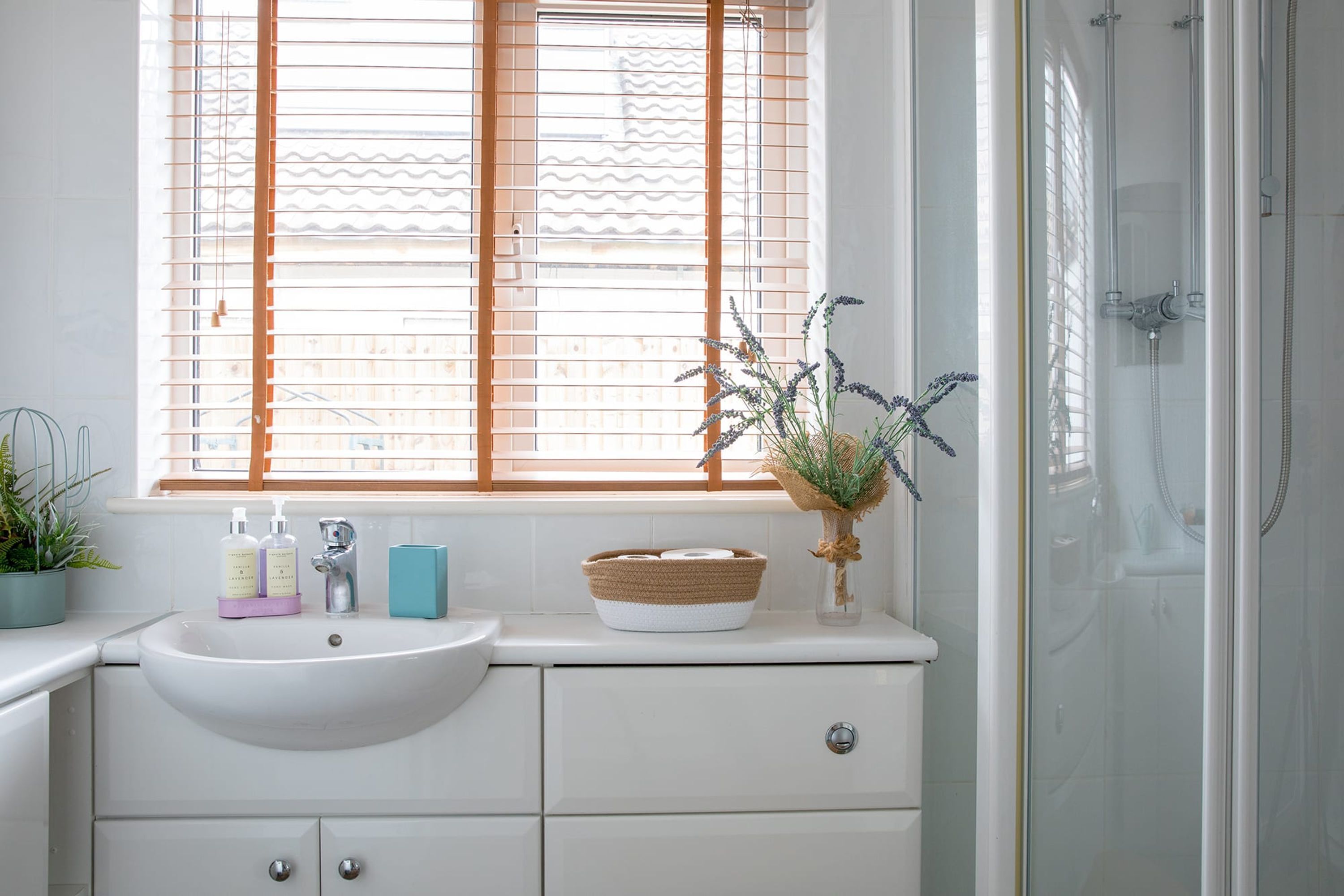 A bathroom with a white sink, wooden window blinds, a shower, and a vase with dried plants.