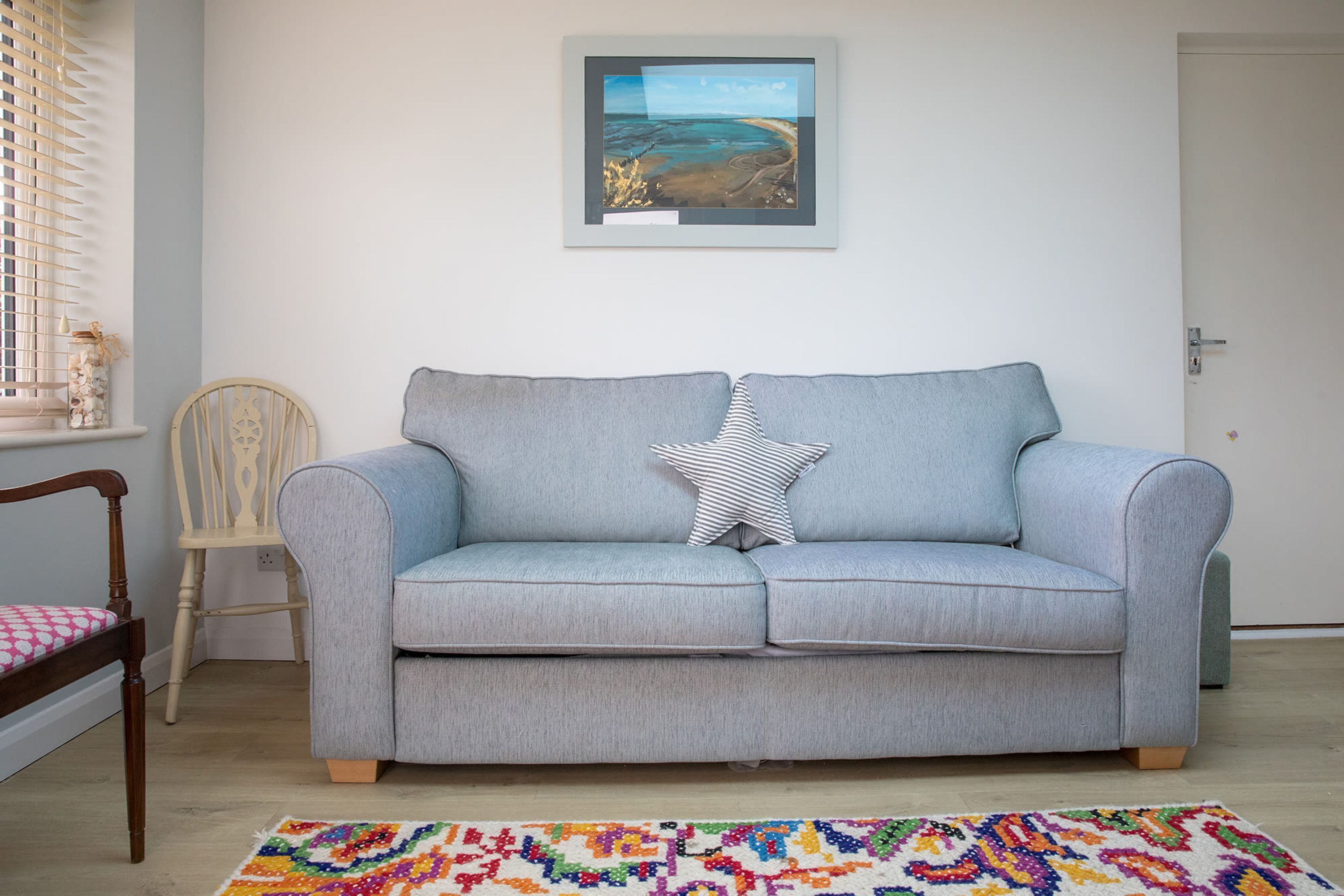 Un sofá gris claro con un cojín en forma de estrella, una silla de madera con un cojín de cuadros, y una alfombra colorida sobre un suelo de madera.