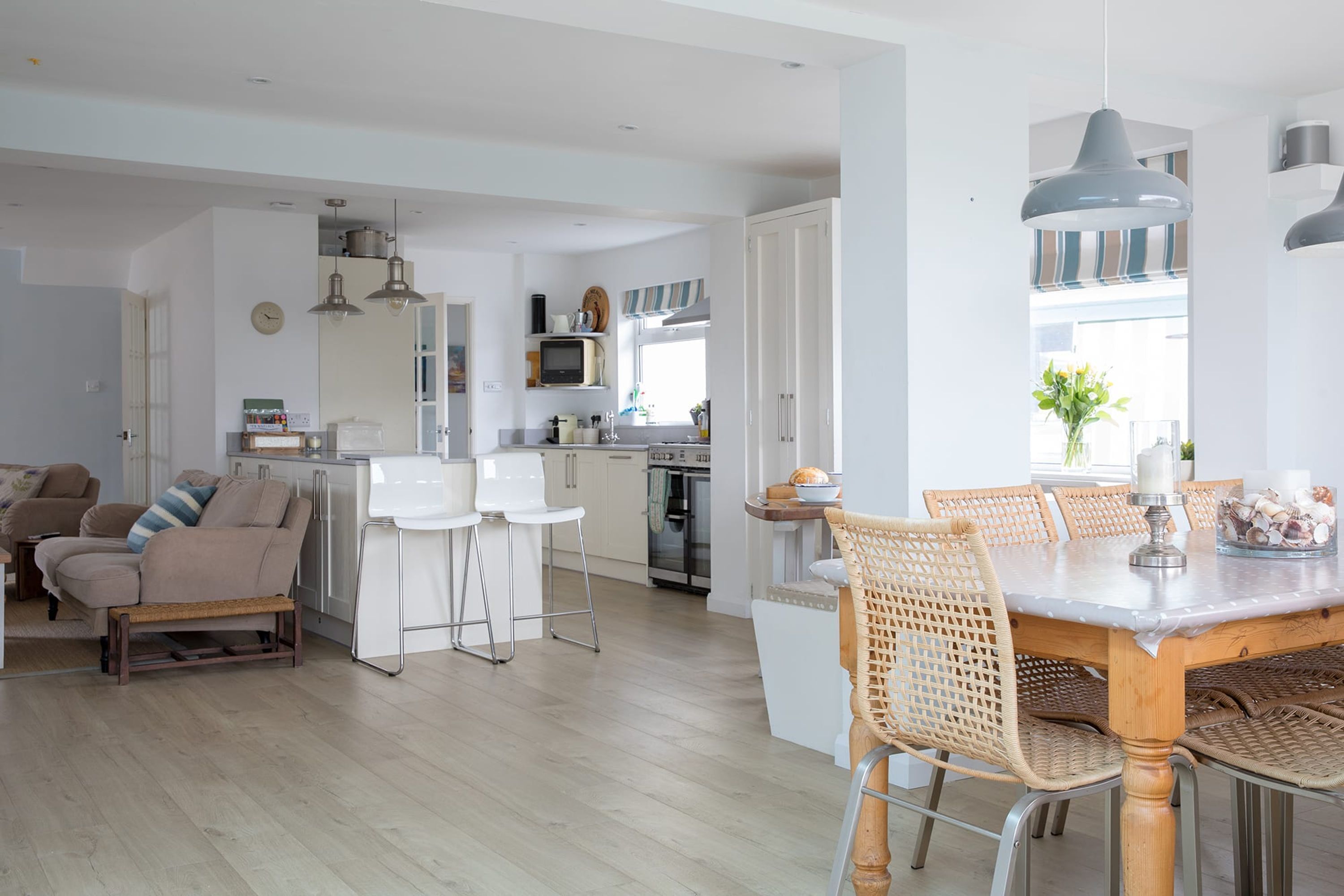 Et moderne køkken med hvide skabe, et spiseområde med et træbord og fletstole, samt en opholdsstue med sofa og sofabord.