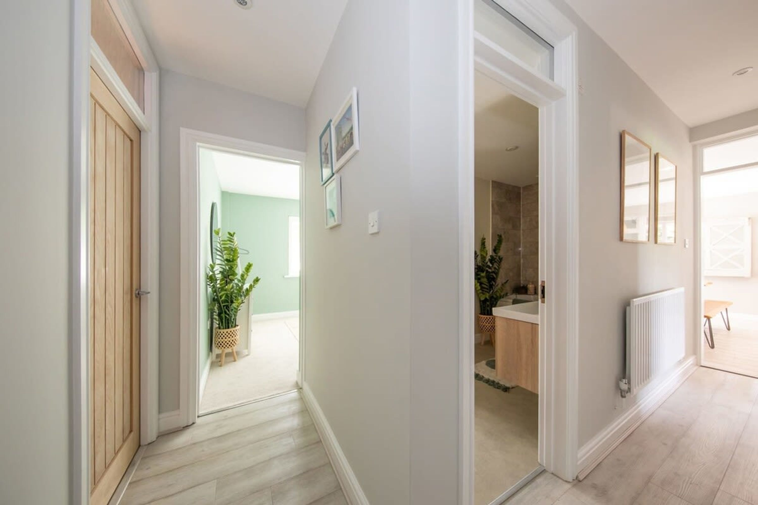 A hallway with a wooden door, a plant, and a view into a bathroom with a sink and a radiator.