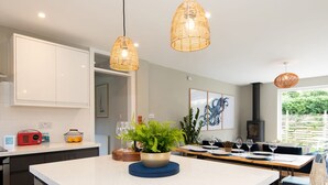 A modern kitchen with a central island, pendant lights, and a dining area with a view of greenery outside. - Bosham 3 BR Beach House With Garden Playhouse (Chichester)
