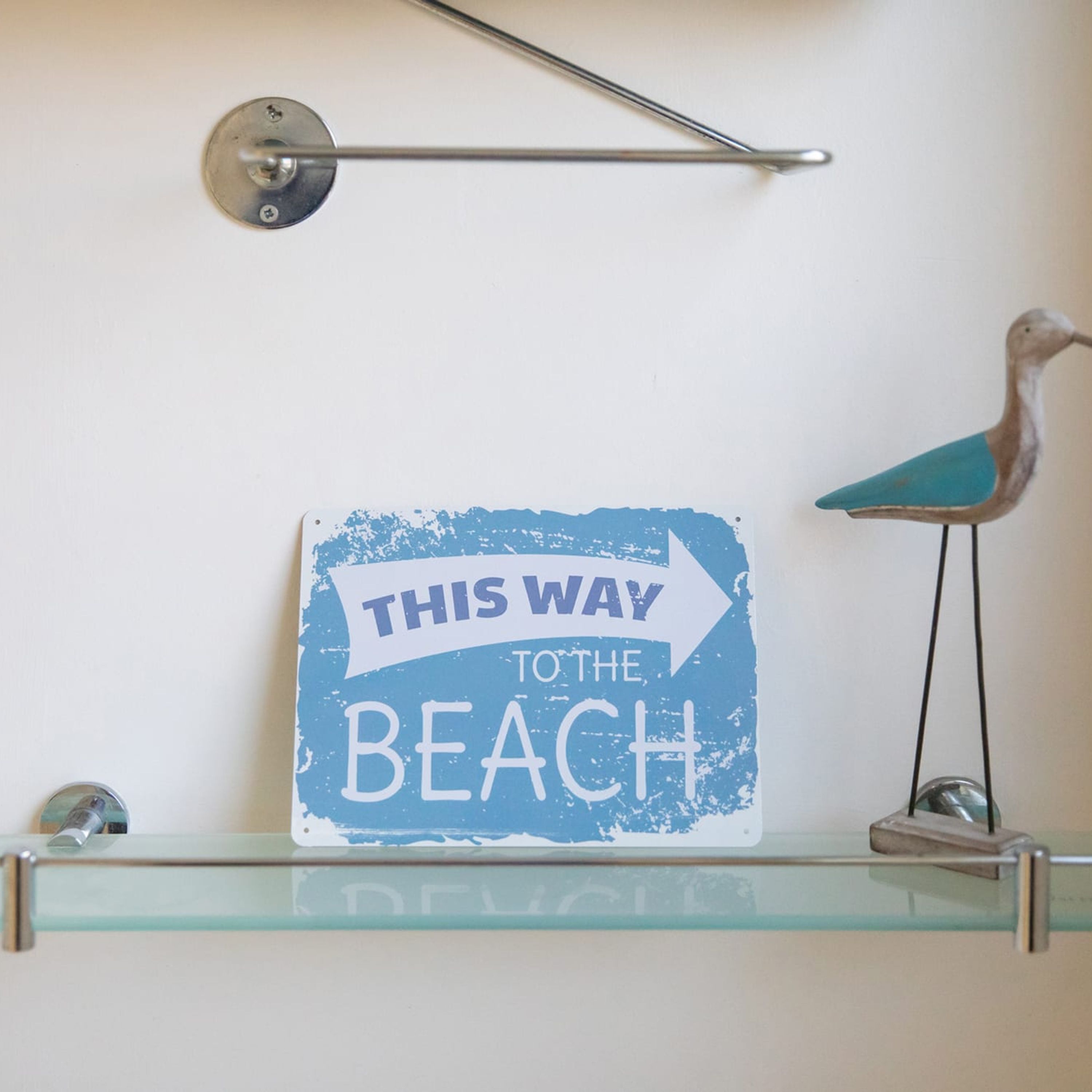 Un letrero en una repisa de vidrio con un soporte metálico que dice: 'THIS WAY TO THE BEACH'.