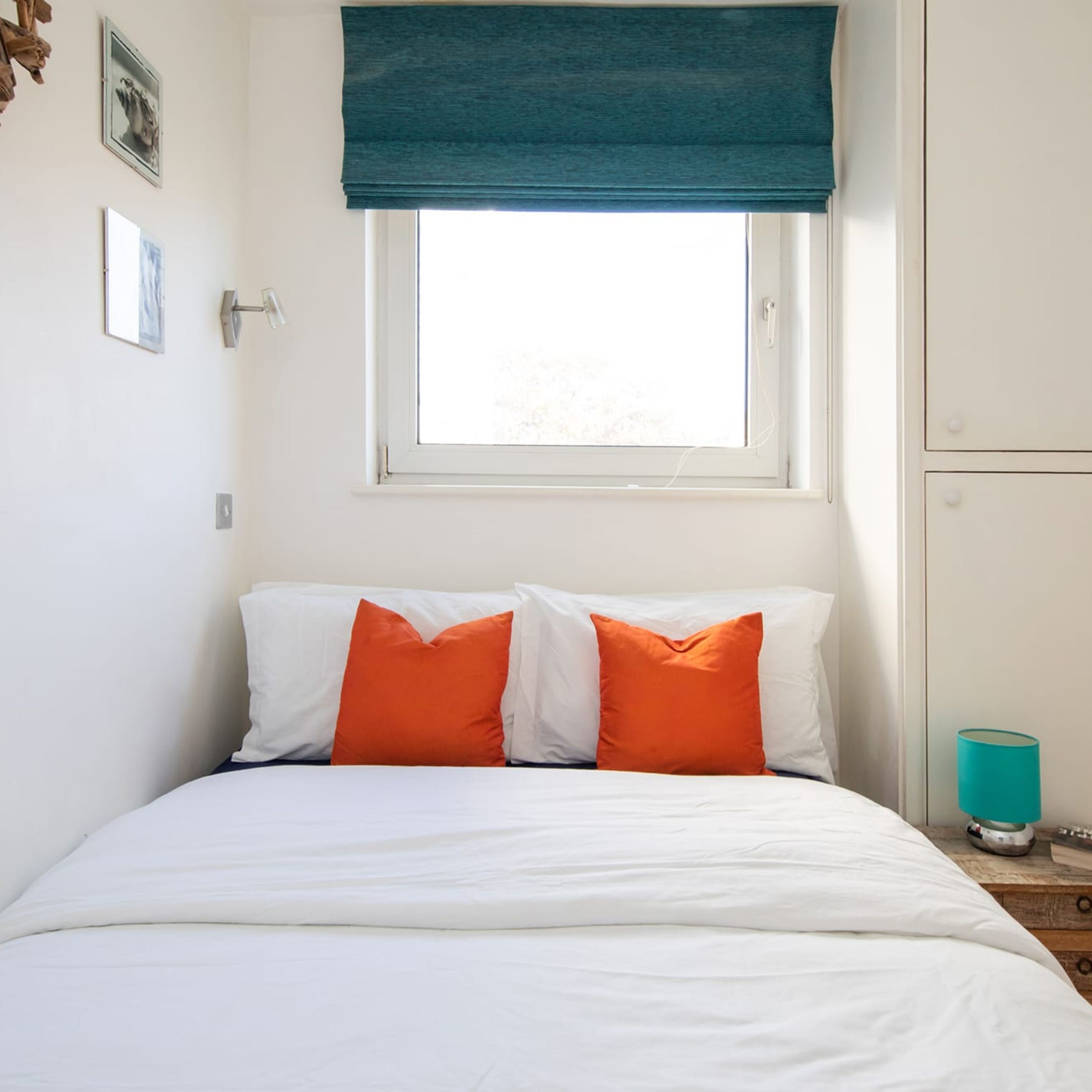 A bedroom with a bed, two pillows, a window with a blue blind, a nightstand with a lamp, and a wall-mounted shelf.
