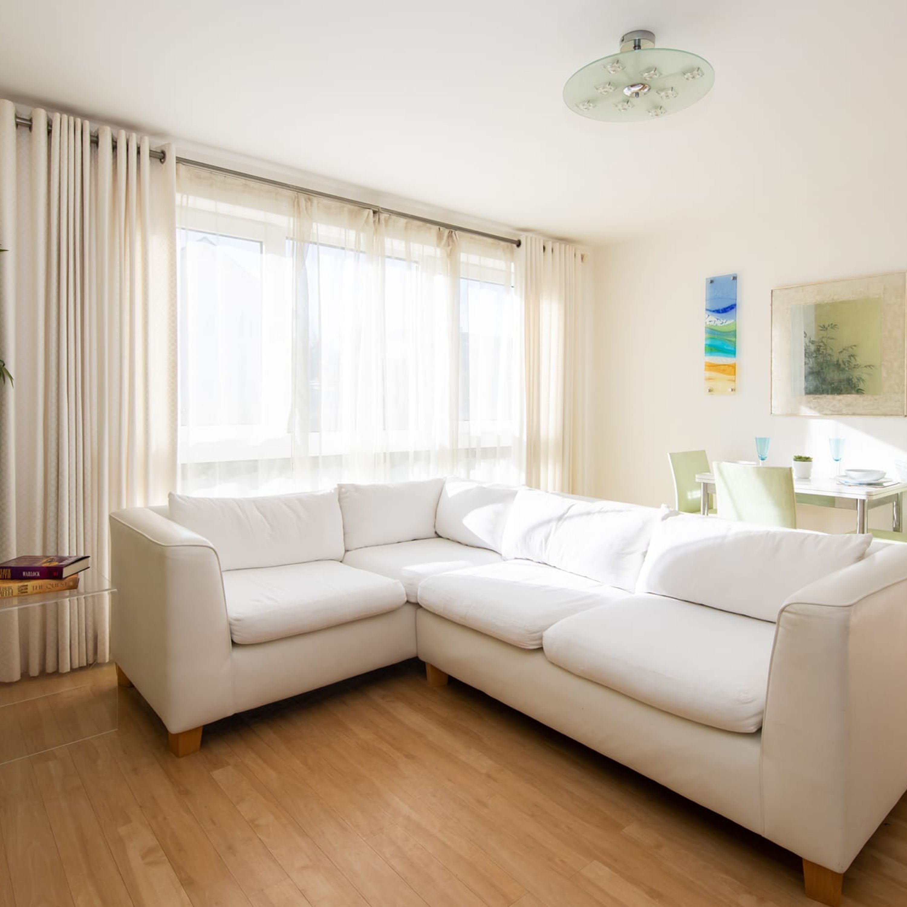 A modern living room with a white sectional sofa, a dining table with chairs, and a large window with sheer curtains.