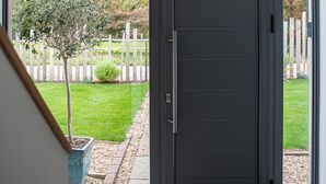 Ein moderner Eingangsbereich mit einer dunkelgrauen Tür, ein kleiner Garten mit Baum und weißem Zaun sowie Blick auf eine grüne Fläche mit Bäumen.