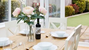 A conservatory with a table set for dining, featuring white chairs, a glass table, and a vase with flowers. - Modern Stylish Seaside Family Home (Chichester)