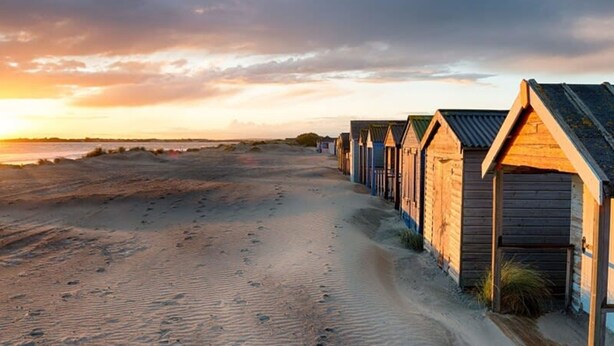 A row of wooden beach huts along a sandy beach with footprints leading to them. - Modern Stylish Seaside Family Home (Chichester)