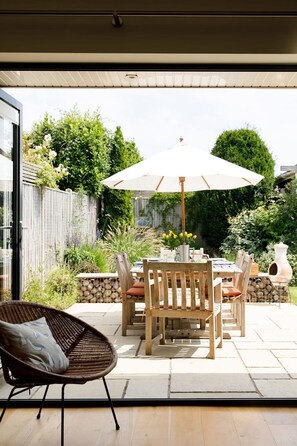 An outdoor patio with a wooden table and chairs, a wicker chair, a white umbrella, and a garden with plants and a fence. - Pretty Coastal Home in Bosham Close to Water (Chichester)