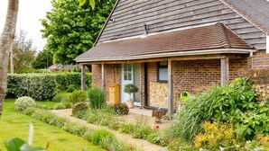 Une maison de plain-pied en briques, dotée d’un toit en pente, d’un porche à l’avant et d’un jardin bien entretenu où poussent diverses plantes, avec un chemin menant à l’entrée.