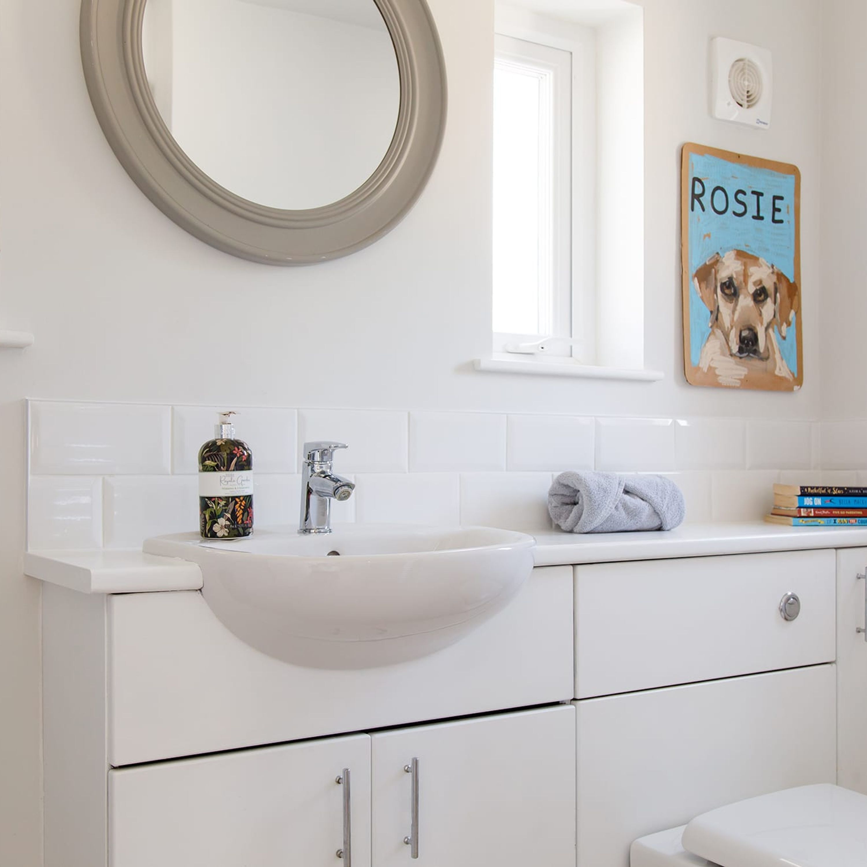 Un baño blanco con un espejo redondo, un lavamanos y un inodoro. En la pared, hay un cuadro con la imagen de un perro que lleva el nombre 'ROSIE'.
