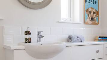 Un baño blanco con un espejo redondo, un lavamanos y un inodoro. En la pared, hay un cuadro con la imagen de un perro que lleva el nombre 'ROSIE'.