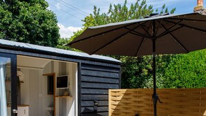 A small, dark wooden shed with a glass door leading to a kitchen area, a round metal table with chairs, and a large umbrella providing shade.