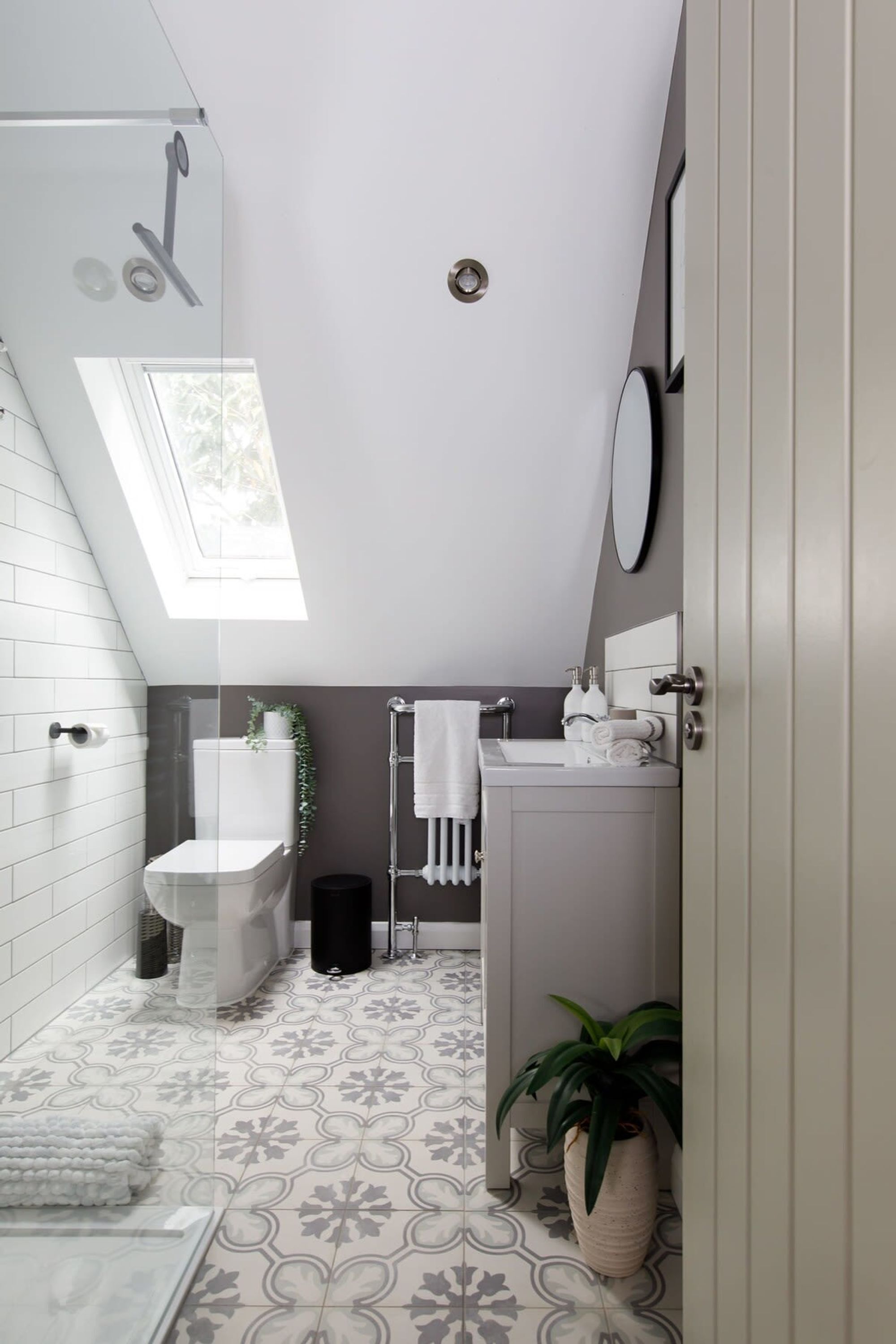 A modern bathroom with a skylight, white tiles, a toilet, a sink, a mirror, and a potted plant.