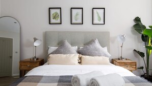 Un lit bien fait, recouvert d’une couvre-lit à carreaux, avec deux lampes de chevet et trois œuvres d’art représentant des feuilles, encadrées et accrochées au mur.