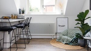 Une pièce moderne et bien éclairée, avec un plafond en pente, un coin repas doté de chaises en métal noir, un radiateur, une grande fenêtre et un tableau encadré accroché au mur.