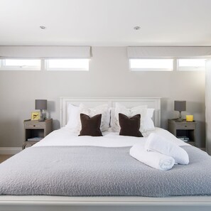 Een net opgemaakt bed met witte lakens en twee bruine kussens, met aan weerszijden nachtkastjes met lampen en een klein tafeltje met een kaars.