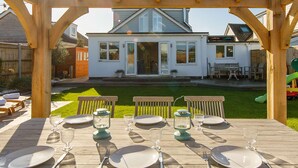Une pergola en bois abritant une table à manger pour quatre personnes, d’où l’on peut admirer une maison blanche et son jardin.