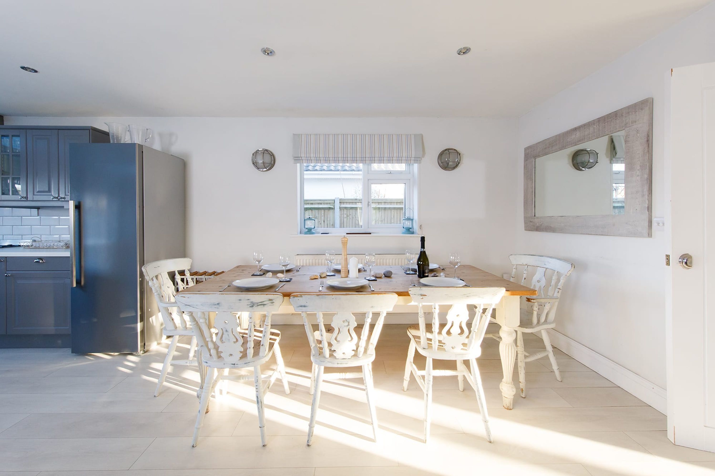 A dining area with a wooden table and white chairs, a refrigerator, and a window with curtains.