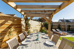 Een houten pergola met een eettafel, omringd door stoelen en een tuin.