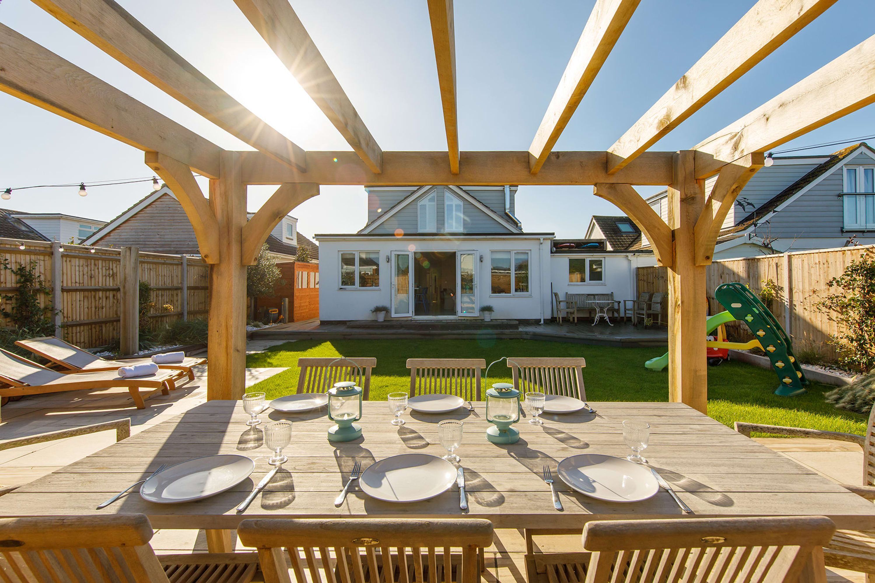 A wooden pergola with a dining table set for six, overlooking a backyard with a house, lawn, and play equipment.