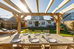 Een houten pergola met een eettafel voor zes personen, met uitzicht op een achtertuin met een huis, gazon en speeltoestellen.