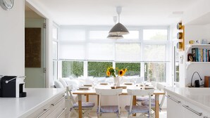 Une cuisine moderne avec des meubles blancs, une table à manger pour quatre personnes et une grande fenêtre donnant sur un paysage verdoyant.