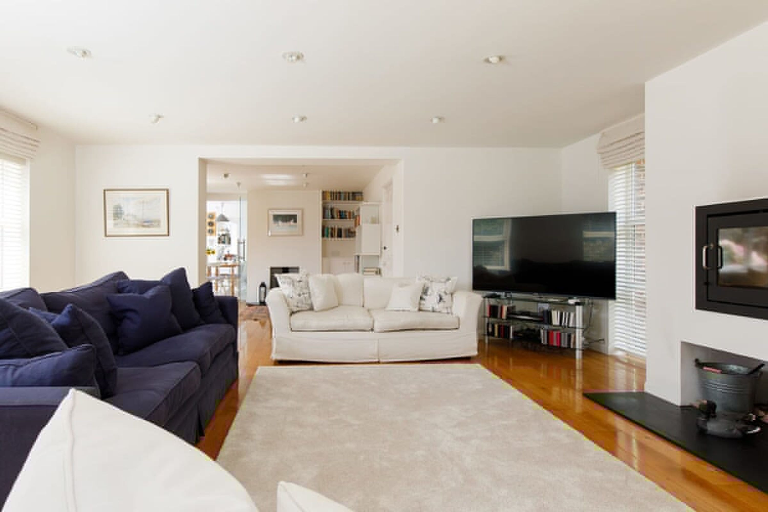 A spacious living room with a large TV, a dark blue sofa, a white sofa, and a beige carpet.