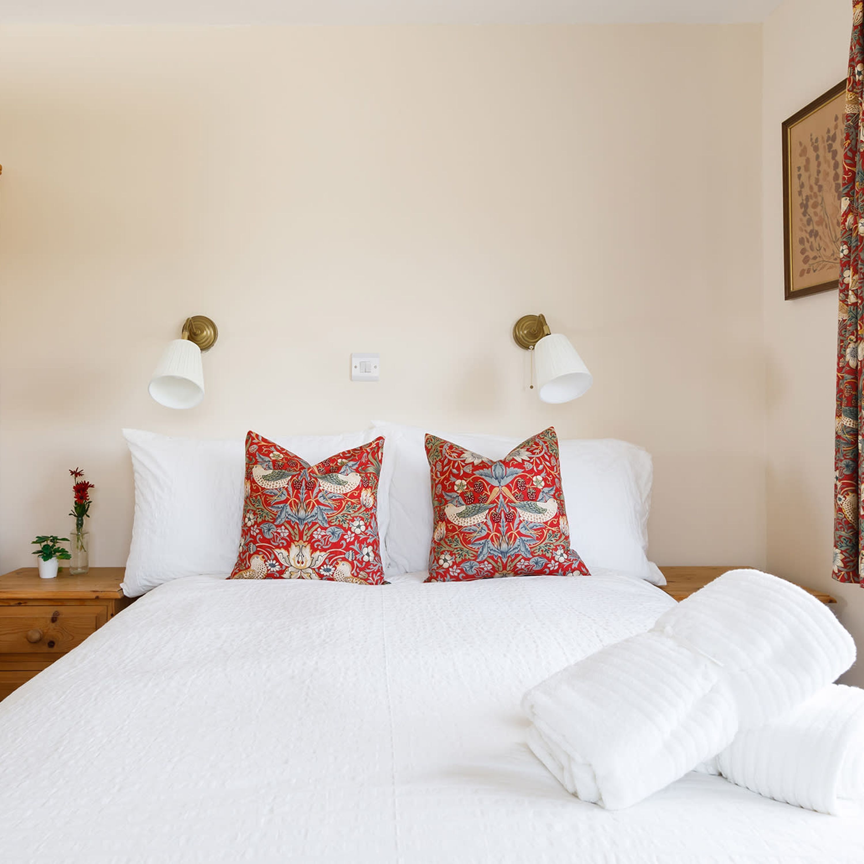A bed with white bedding, two decorative pillows with a red floral pattern, two wall-mounted lamps, a small bedside table with a vase of flowers, and a framed picture on the wall.
