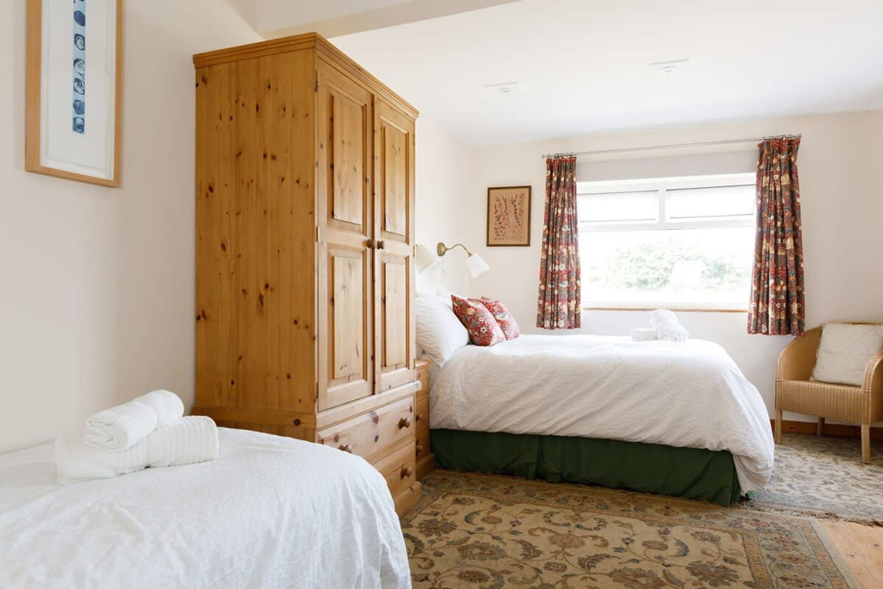 A bedroom with two beds, a wooden wardrobe, a patterned rug, and a window with curtains.