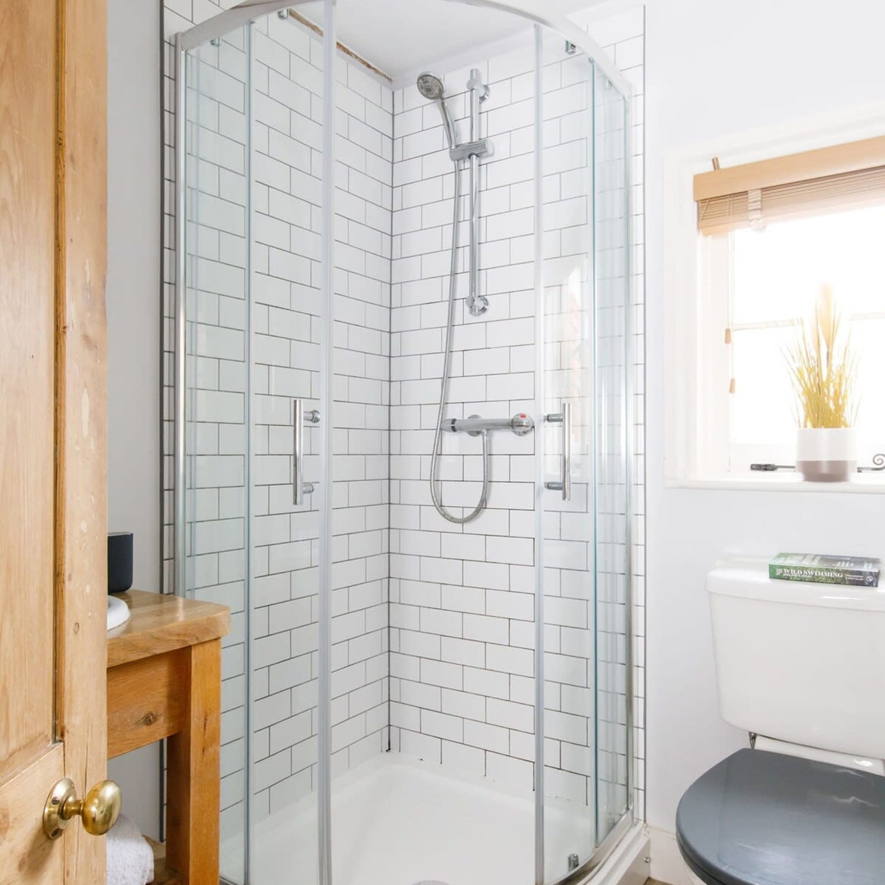 Une salle de bain d’angle avec une douche à parois de verre, des murs recouverts de carreaux blancs, une porte en bois et une fenêtre agrémentée d’une plante.