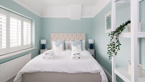 A bedroom with a large bed, two bedside tables, a window with blinds, and a plant on a white shelf. - Bright Modern 4 br House 100 Metres From Beach (Chichester)