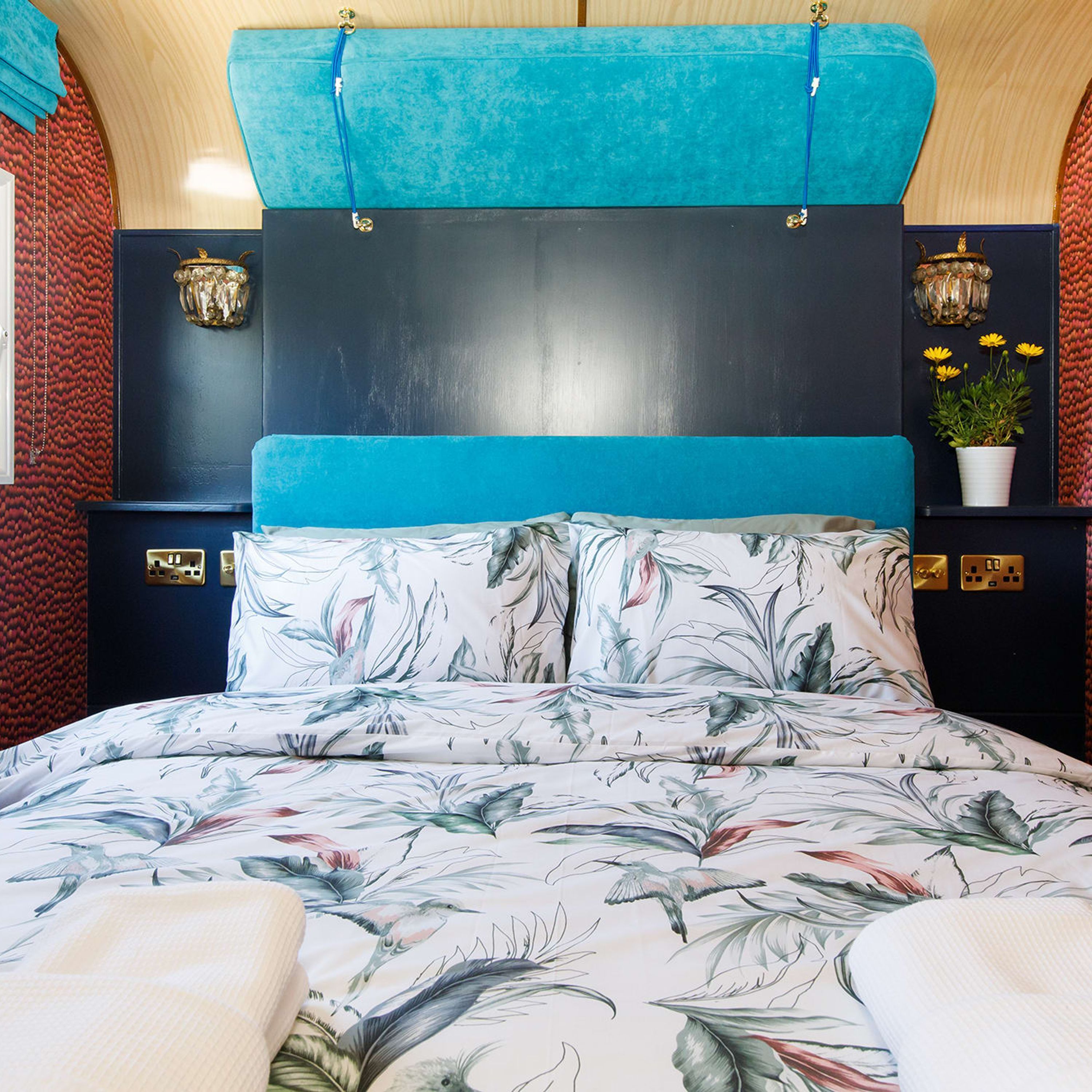A bedroom with a bed featuring a floral print, a teal upholstered headboard, and a dark wall with decorative wall sconces.