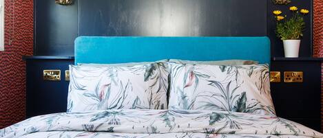 A bedroom with a bed featuring a floral print, a teal upholstered headboard, and a dark wall with decorative wall sconces.