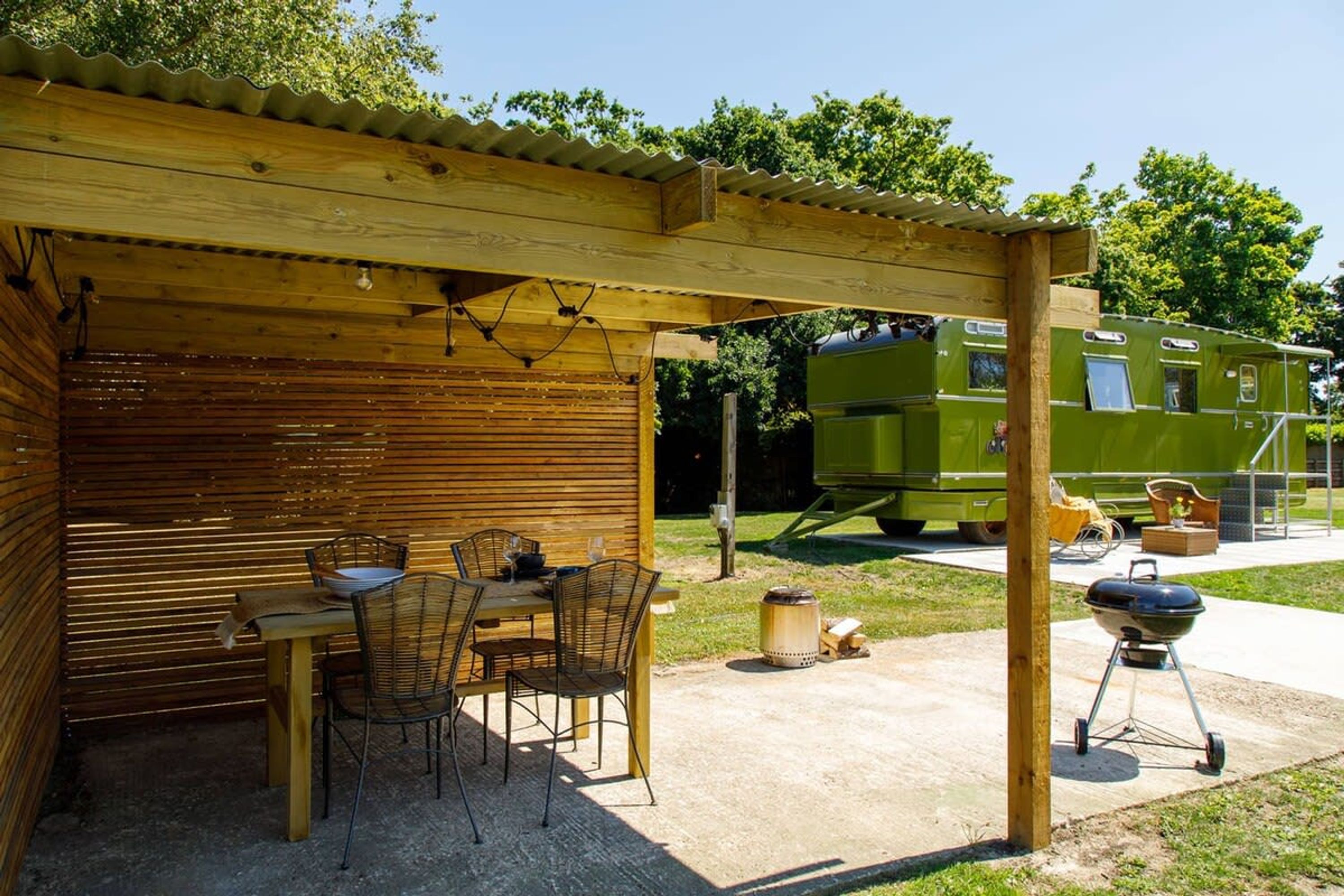 Une terrasse extérieure couverte, avec une table et des chaises en bois, un barbecue et, au fond, un camping-car vert.