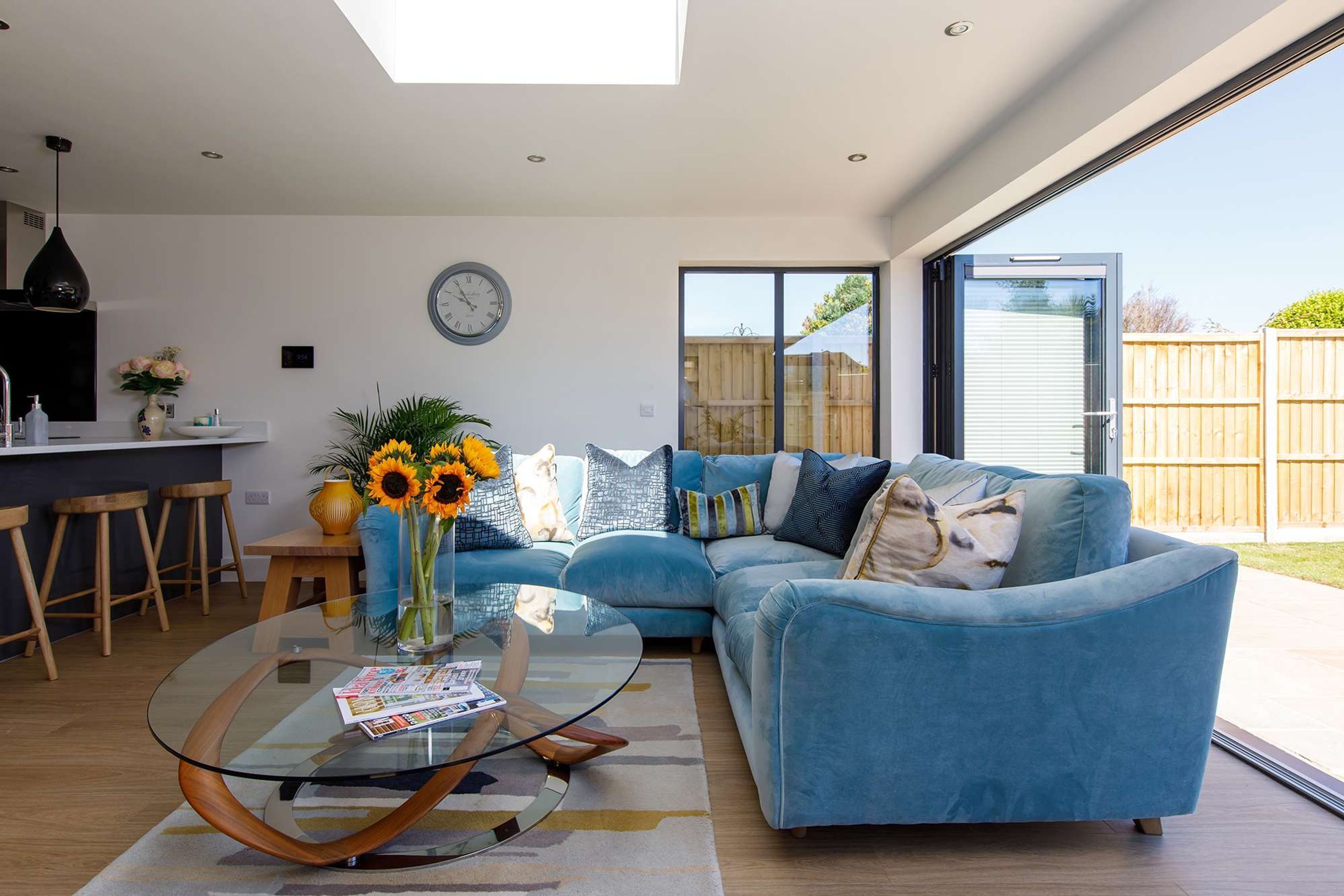 A modern living room with a glass coffee table, a blue sofa, and a dining area with wooden stools.