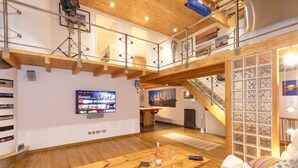 A spacious loft-style living area with a wooden floor, a large wooden table, a sofa, and a TV mounted on the wall. - Stylist Split Level 2 Bed With Parking (Chichester)