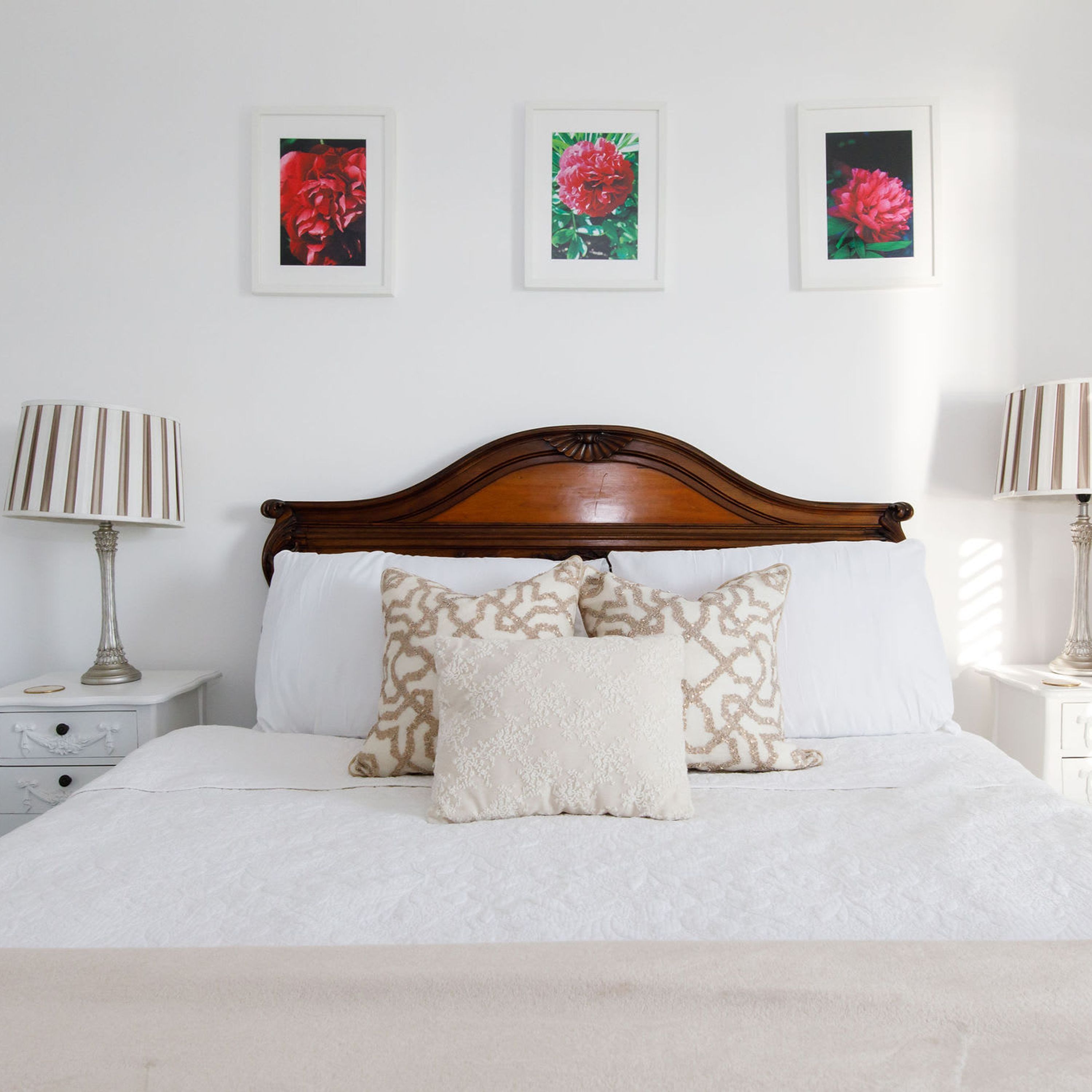 A neatly made bed with a wooden headboard, white bedding, and patterned pillows. There are three framed floral pictures on the wall above the bed.
