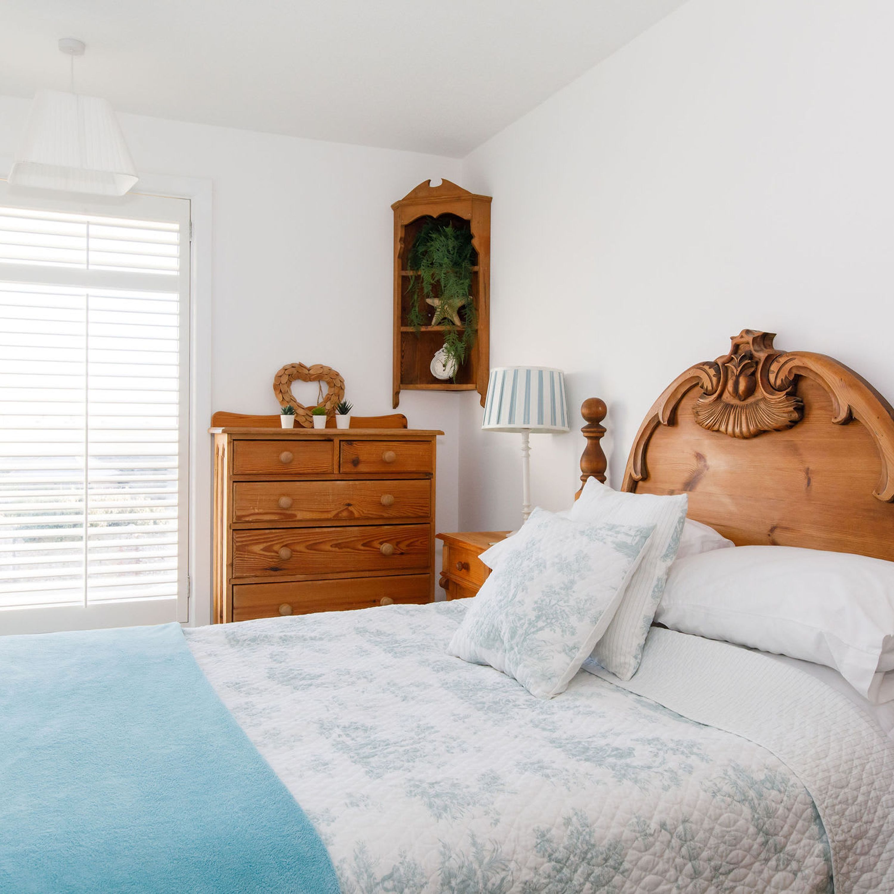 A bedroom with a wooden dresser, a nightstand, a bed with floral bedding, a heart-shaped wall decoration, and a plant on a shelf.