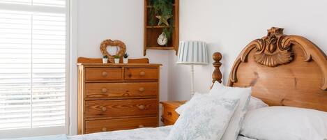 A bedroom with a wooden dresser, a nightstand, a bed with floral bedding, a heart-shaped wall decoration, and a plant on a shelf.