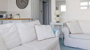 Un salon moderne aux couleurs claires, avec une grande horloge murale, une petite table avec une lampe et une porte-fenêtre donnant sur une autre pièce.
