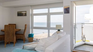 Une pièce avec un canapé blanc, un fauteuil en osier, une petite table et une vue sur la plage, visible à travers de grandes fenêtres.