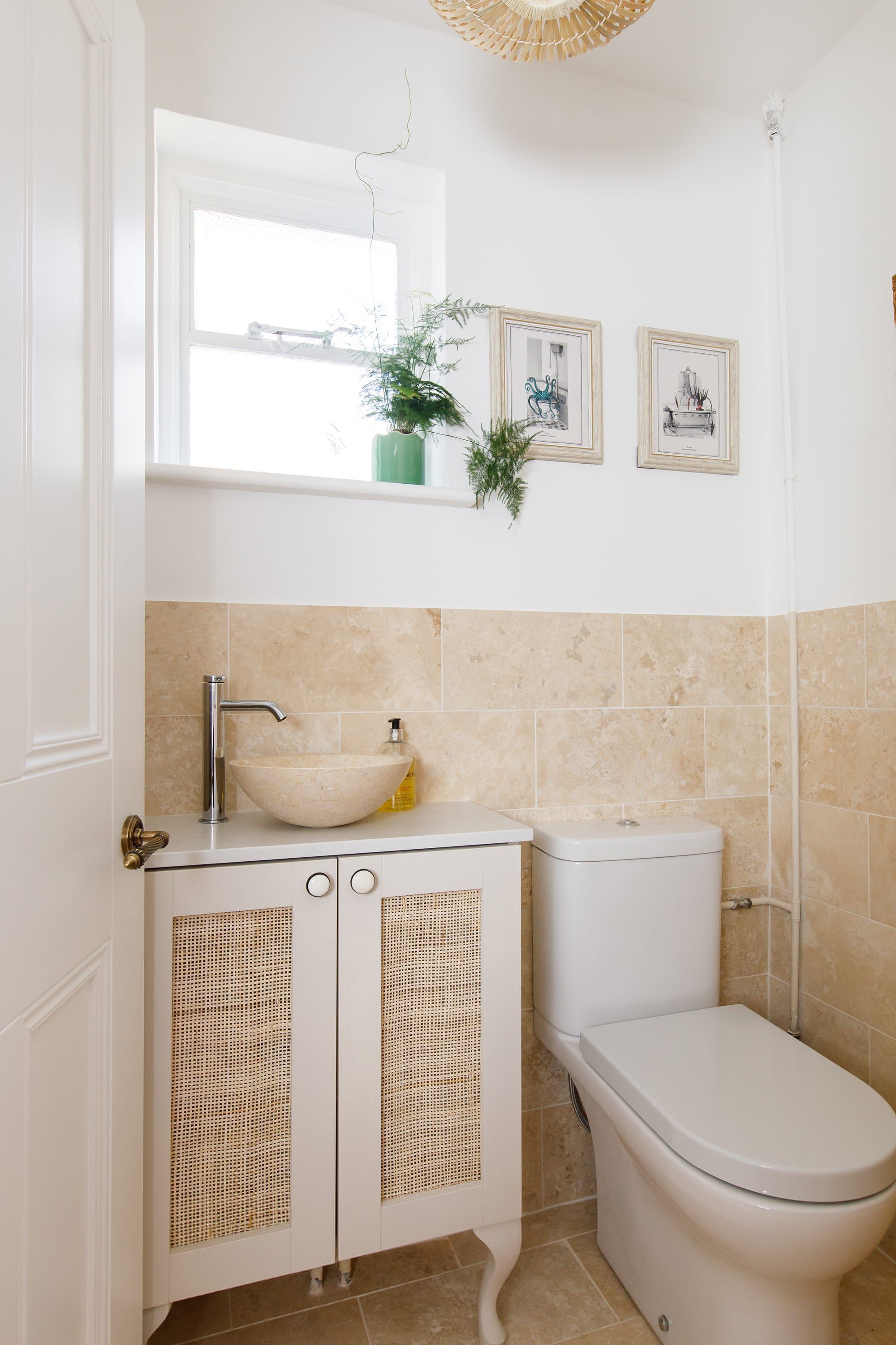A bathroom with a white sink, toilet, and wicker cabinets. There are framed pictures on the wall and a plant on the windowsill.