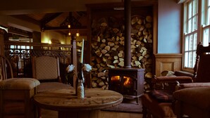 A cozy room with a wooden floor, a round table with a vase, chairs, a fireplace with a log stack, and a window. - Serene Riverside 6BR Luxury Retreat in Curdridge (Southampton)